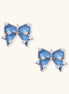 Pair of bow-shaped stud earrings featuring blue denim fabric with white stitching and gold-plated crystal accents