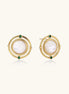 Close-up of gold-plated circular earrings with white mother-of-pearl centers and green zirconia accents on cream background.