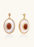 Gold-plated drop earrings with oval mother-of-pearl stones, central red gemstones, and crystal accents on white background.