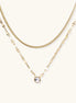 Two layered 14K gold plated necklaces - herringbone chain and delicate chain with circular crystal pendant on beige background.