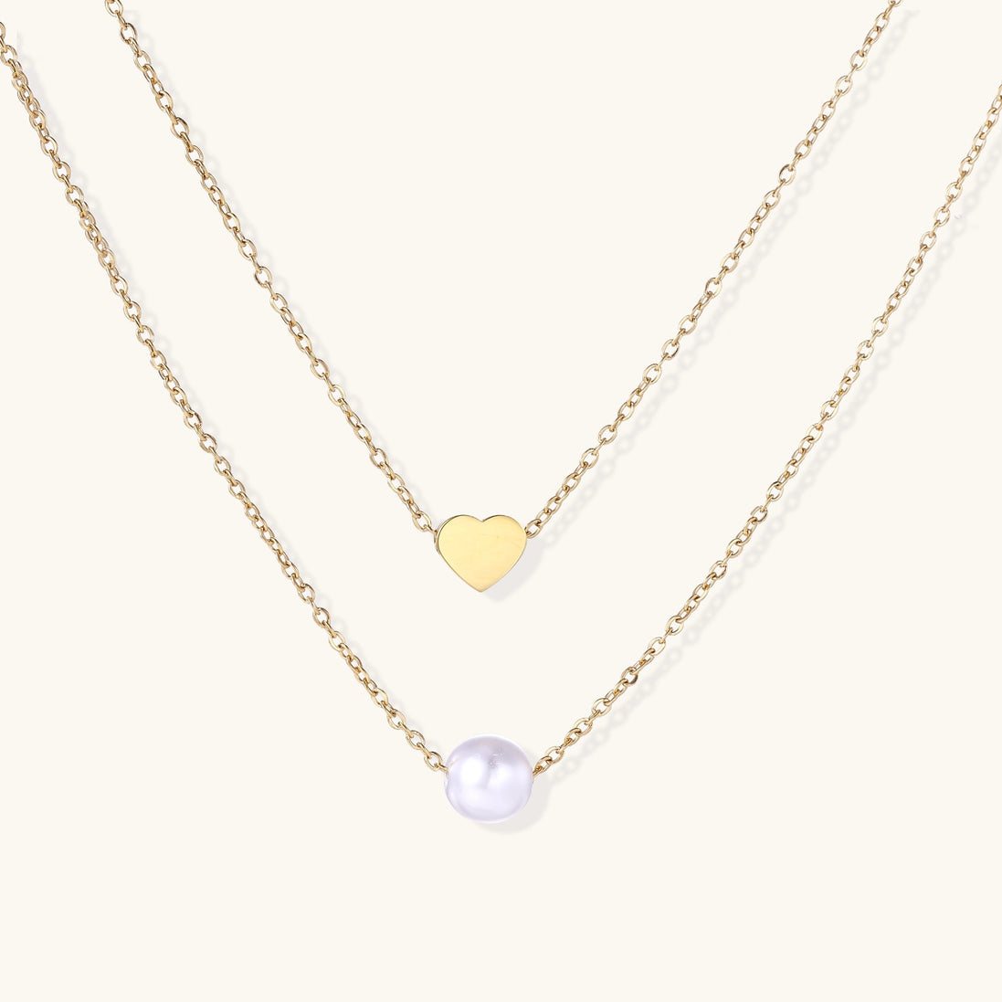 Two delicate gold-plated chains layered together, one with small heart pendant, another with cream pearl bead
