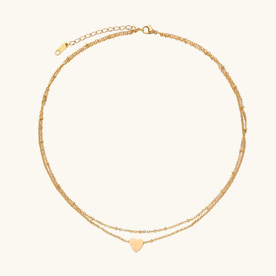 Two-layer gold-plated stainless steel necklace with small heart pendant displayed on beige background