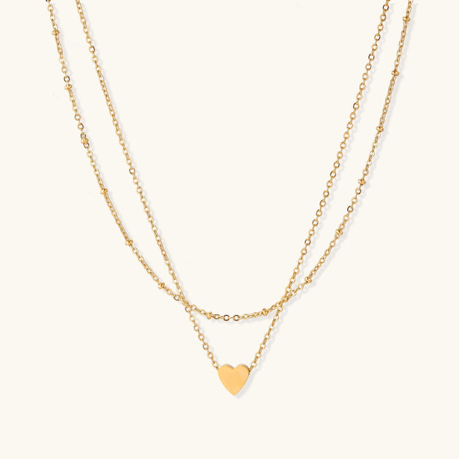 Two-layer gold-plated stainless steel necklace with small heart pendant displayed on beige background