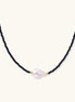 Delicate necklace with black blue sandstone beads and baroque pearl center on white background with gold accents.