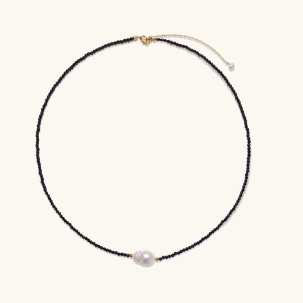 Delicate necklace with black blue sandstone beads and baroque pearl center on white background with gold accents.