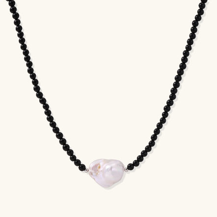 Beaded black agate necklace with iridescent baroque freshwater pearl centerpiece on cream background.